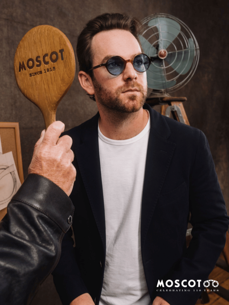 Zack Moscot wearing sunglasses is holding a wooden paddle with the word moscot on it.