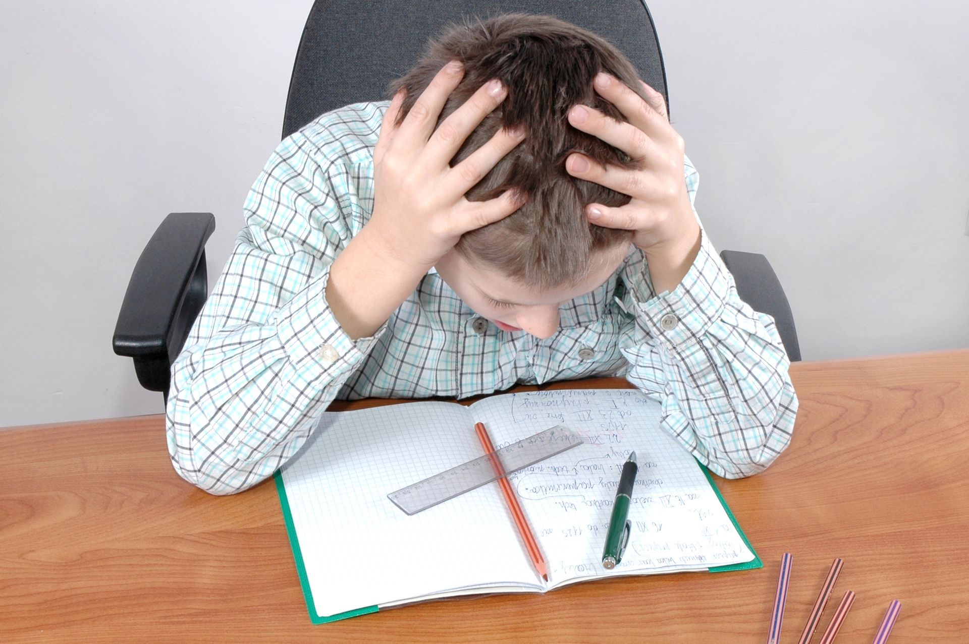 Boy stressed, holding head in hands, over a notebook with a ruler and pencils on a desk.