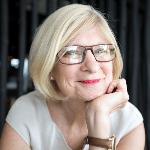 Woman with  blonde hair and glasses, resting chin on hand, smiling. 