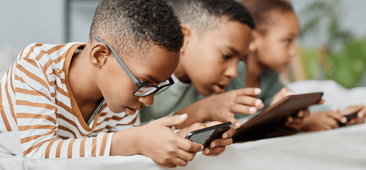 Three children engrossed in devices, lying down. One with glasses holds a phone; others use tablets.