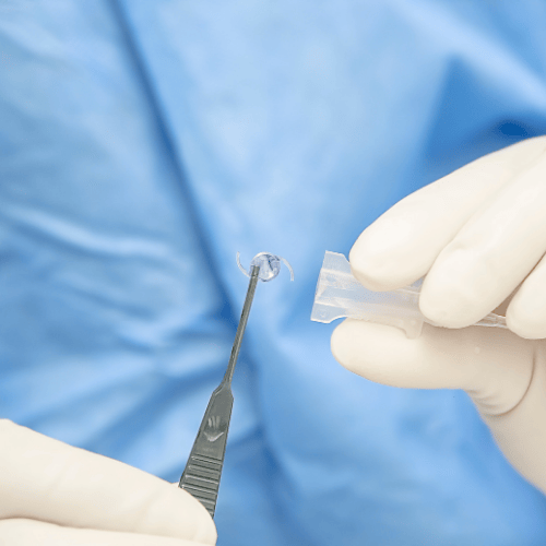 Surgeon holding an intraocular lens with forceps, next to its plastic case, in a sterile setting.