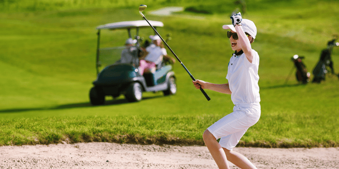 A young boy is hitting a golf ball out of a bunker on a golf course, wearing sport sunglasses.