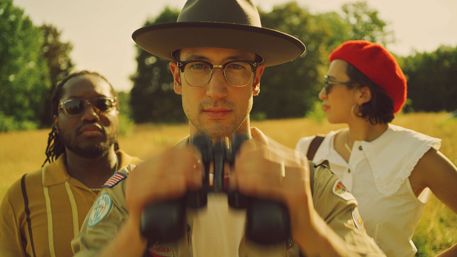 A man wearing glasses and a hat is looking through binoculars in a field.