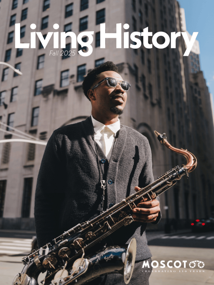 A Black man in sunglasses holding a saxophone, standing on a city street with a building in the background.