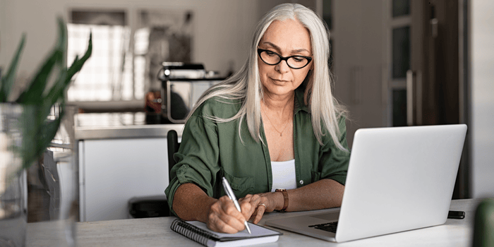 Woman with long white hair and glasses writing in a notebook, using a laptop in a well-lit room.
