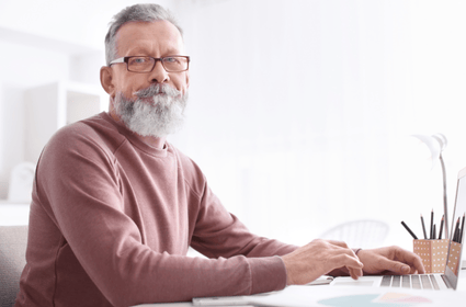 Older man with glasses and gray beard working on a laptop in a light-filled room.