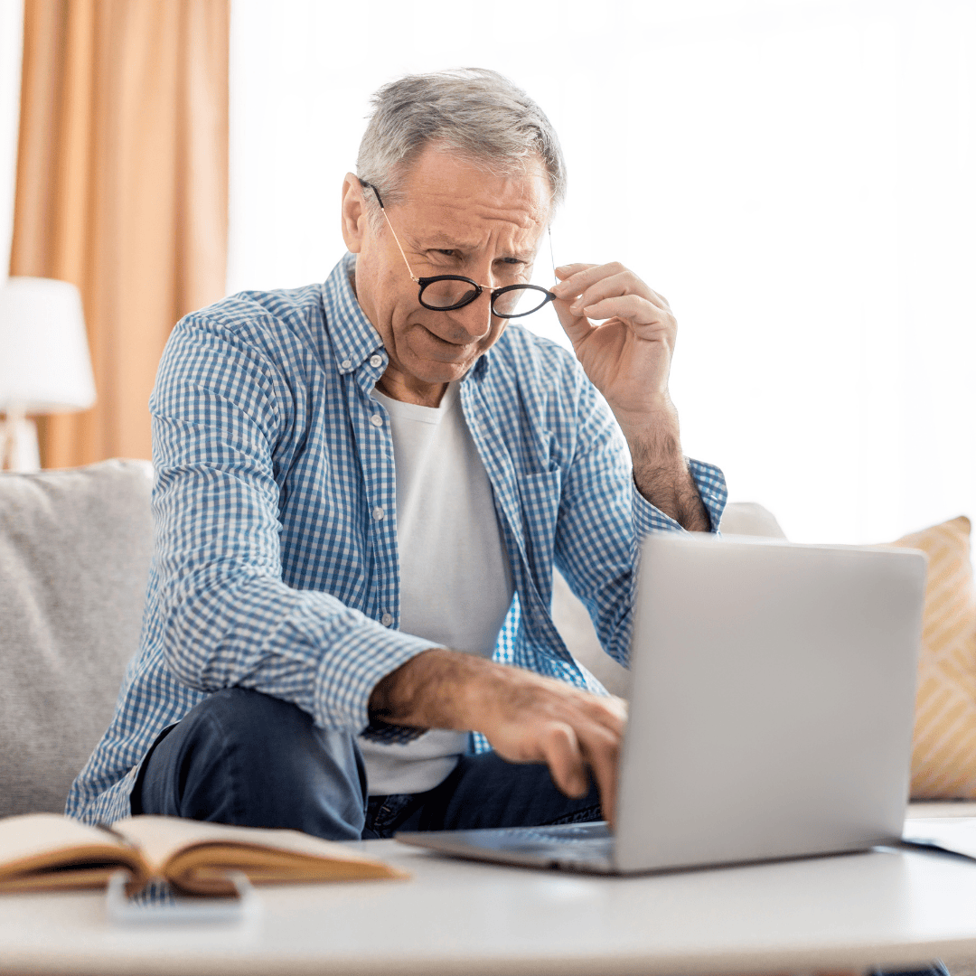 Older man with glasses looking at a laptop, sitting on a couch.