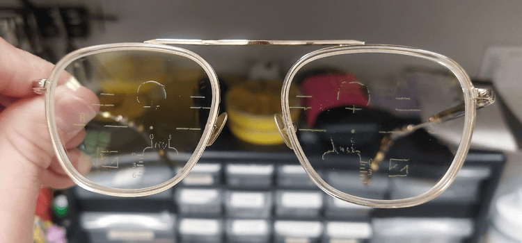 Gold eyeglasses with visible progressives lines, held against a blurred background of objects.