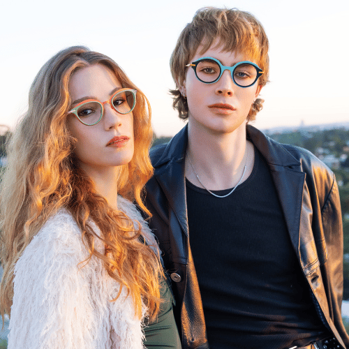 Two people with glasses posing outdoors in warm sunlight, one in a white fuzzy jacket and one in a black jacket.