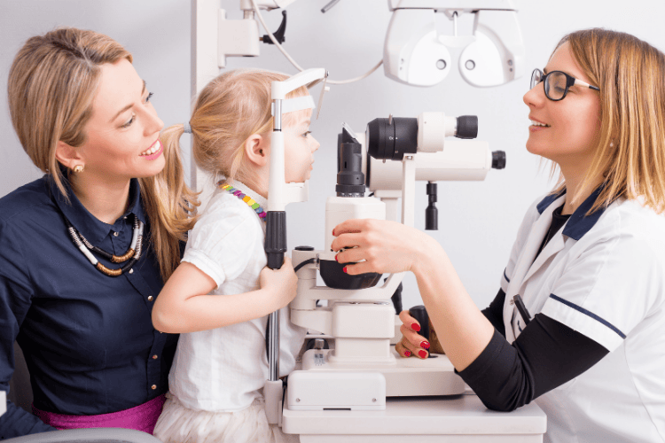 Child receiving eye exam with doctor, mother present. White medical office.