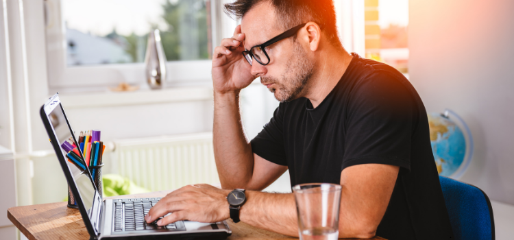 A man with glasses and a black shirt looking stressed while working on a laptop.