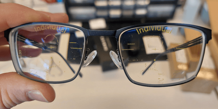 A pair of eyeglasses with rectangular lenses, held in a hand. The glasses have a dark frame.