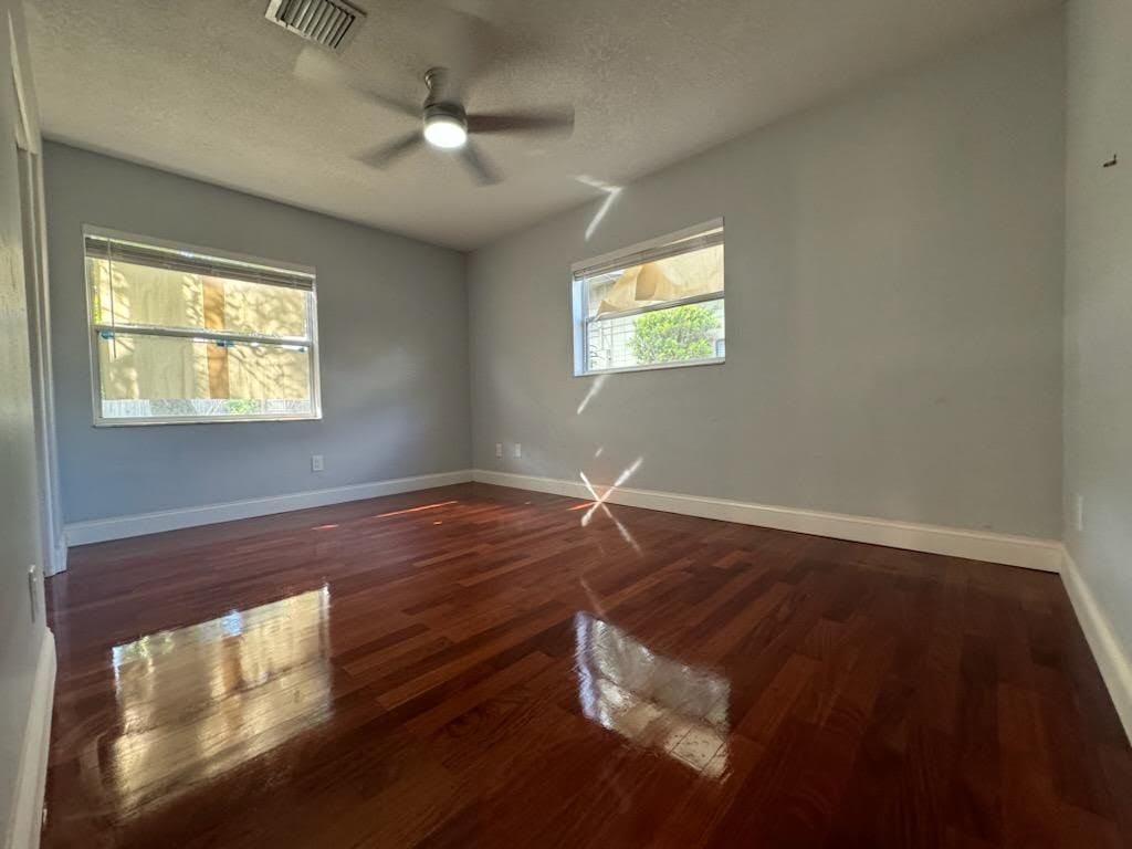 Empty room with gray walls, glossy hardwood floor, ceiling fan, and two windows with white trim.