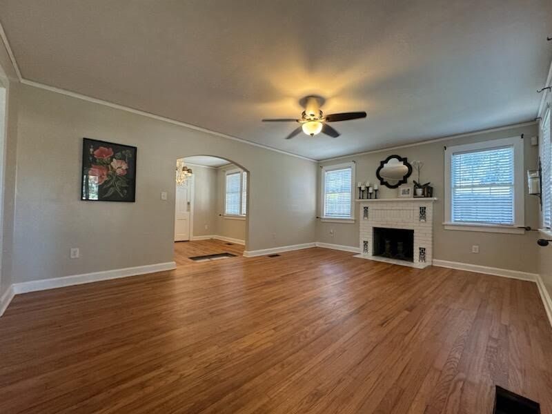 Empty living room with hardwood floors, white walls, fireplace, ceiling fan, and windows.