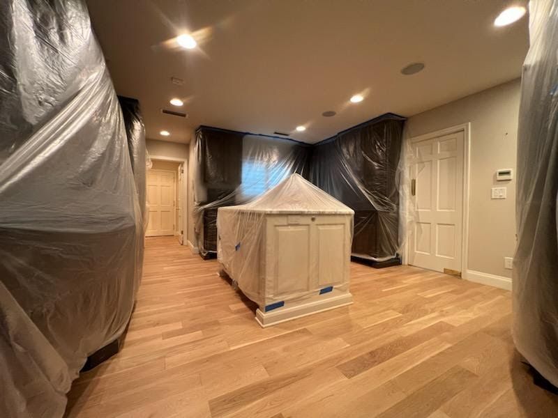Living room under renovation, with plastic-covered furniture and a small covered cabinet on hardwood floors.