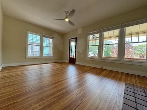 Empty living room with hardwood floors, beige walls, large windows, and a ceiling fan