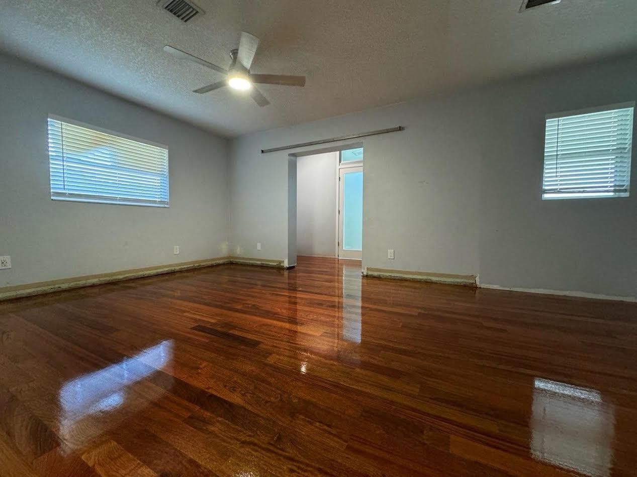 Empty room with polished hardwood floors, white walls, windows, and an open doorway to another room