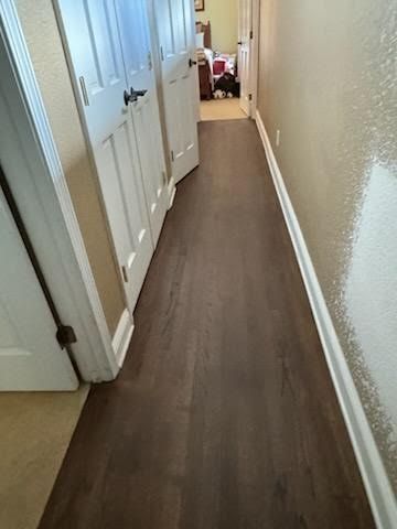 Narrow hallway with white doors, beige walls, and brown wood flooring leading to a room at the end