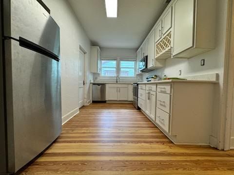 Narrow kitchen with stainless fridge, white cabinets, wood floor, and a window over the sink.