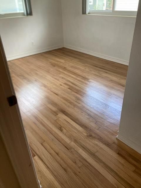 Empty room with light wood floors, white walls, and two windows letting in sunlight.