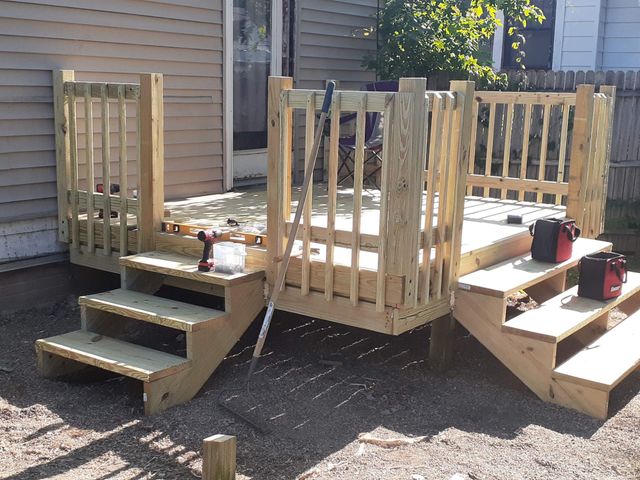 Person kneeling on a wooden deck frame, using tools to build a backyard deck beside a house.