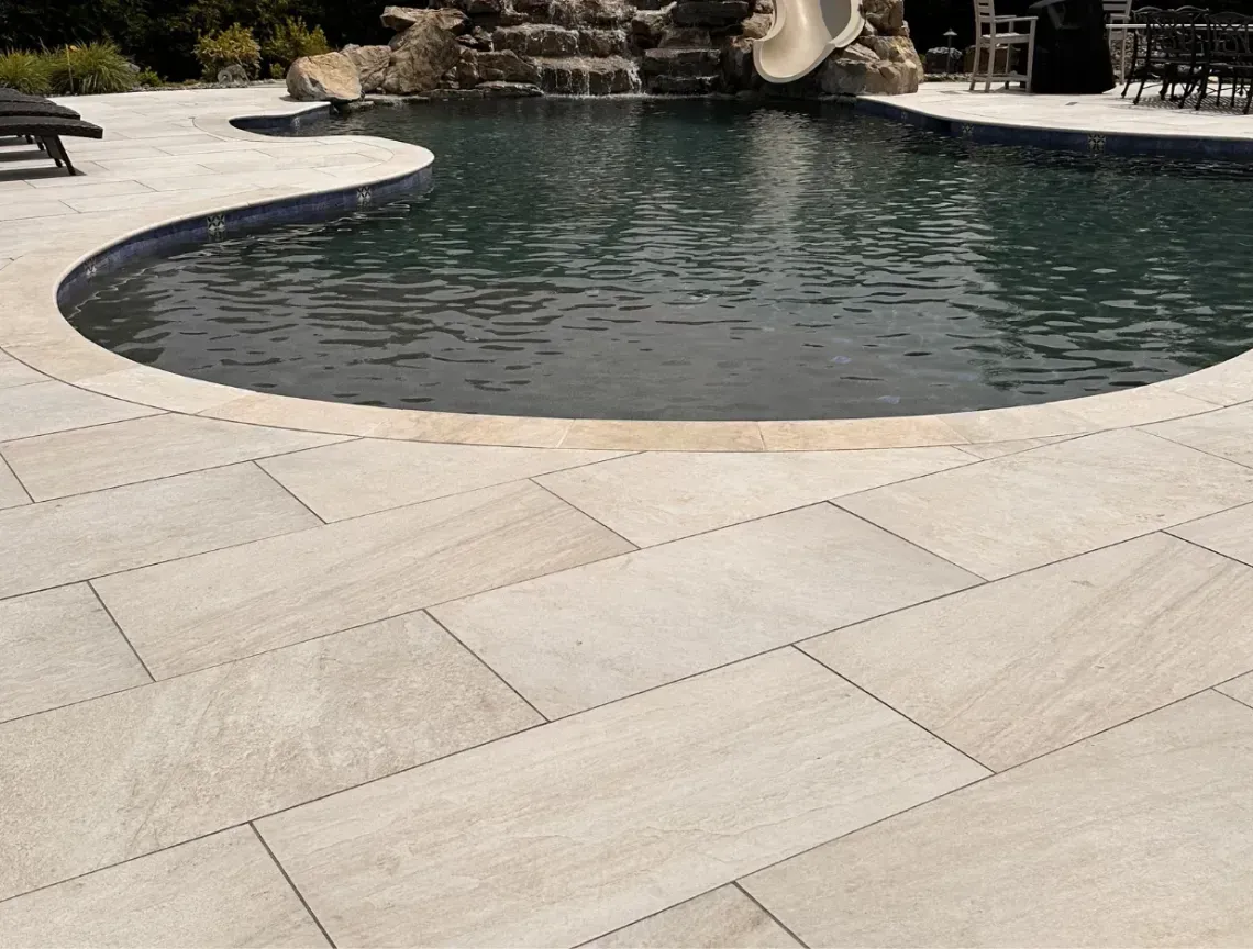 Curved stone patio beside a backyard pool with dark water and rock waterfall feature
