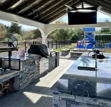 Covered outdoor picnic area with stone tables, black benches, and a grill overlooking a fenced park.