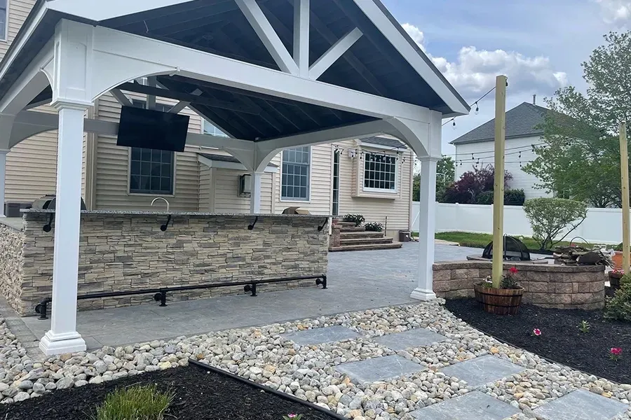 Covered stone patio beside a beige house with white posts, a hanging chair, and landscaped rock path