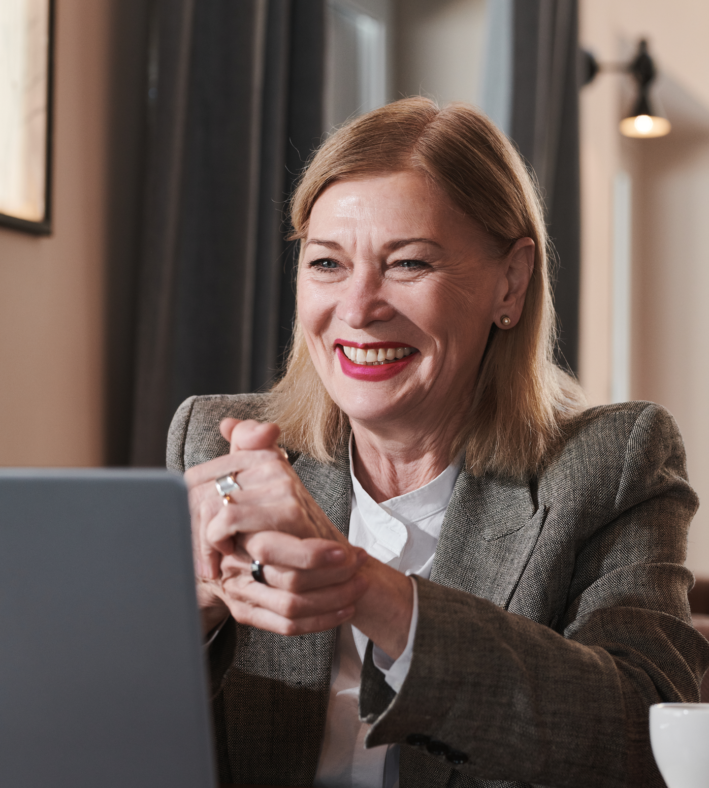 Woman in blazer smiles while using a laptop. She's indoors, near a window, hands clasped.