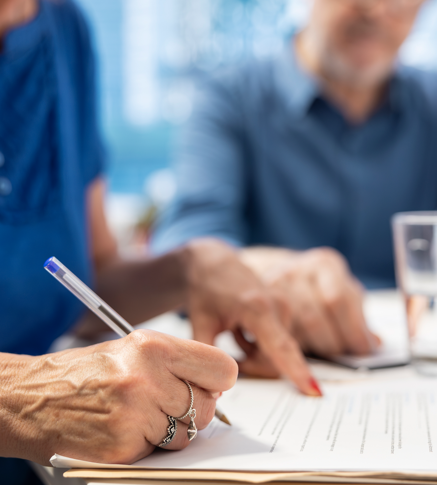 Person signing a document, another person points to the document. Both are seated at a table.