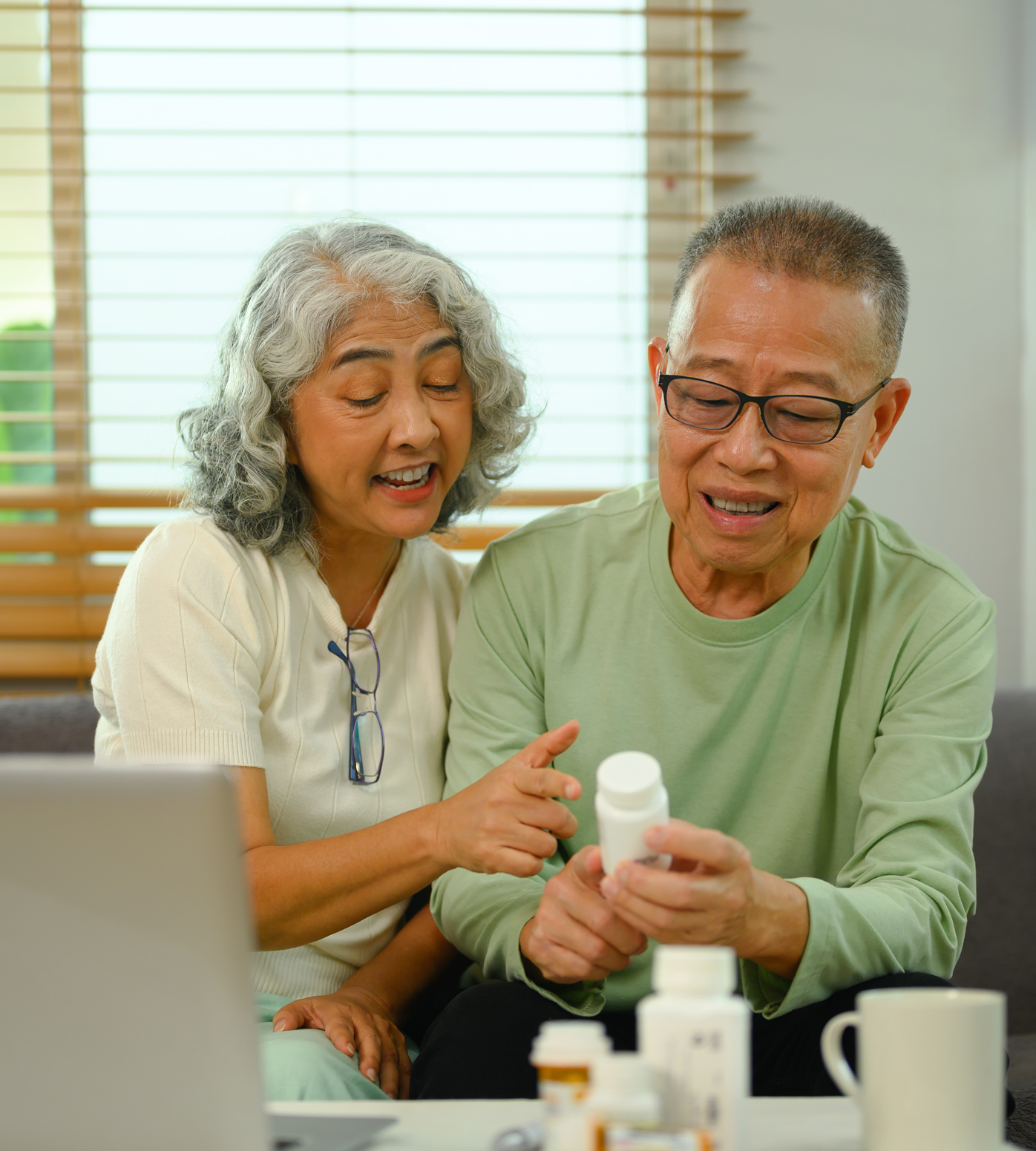 Elderly couple reviewing medicine bottle while using laptop; indoors, light colors, focused.