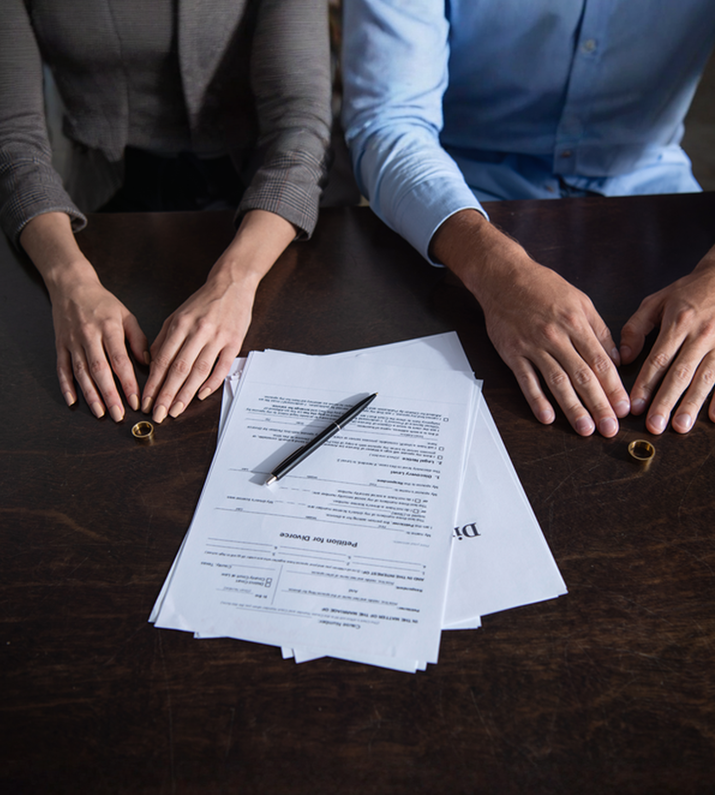 Couple at a table with divorce papers and wedding rings, hands apart.