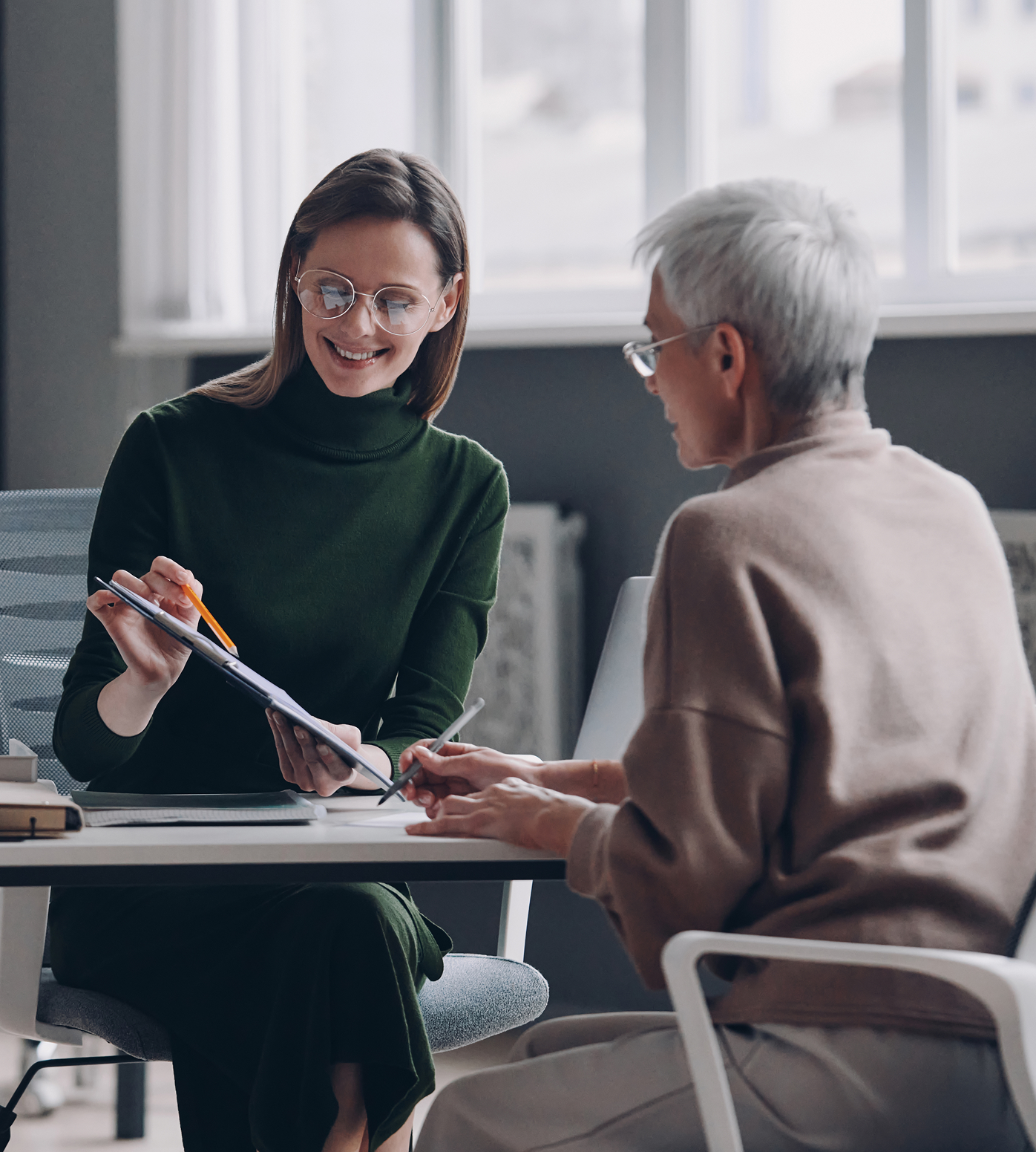 Woman in green dress showing document to older woman at desk.