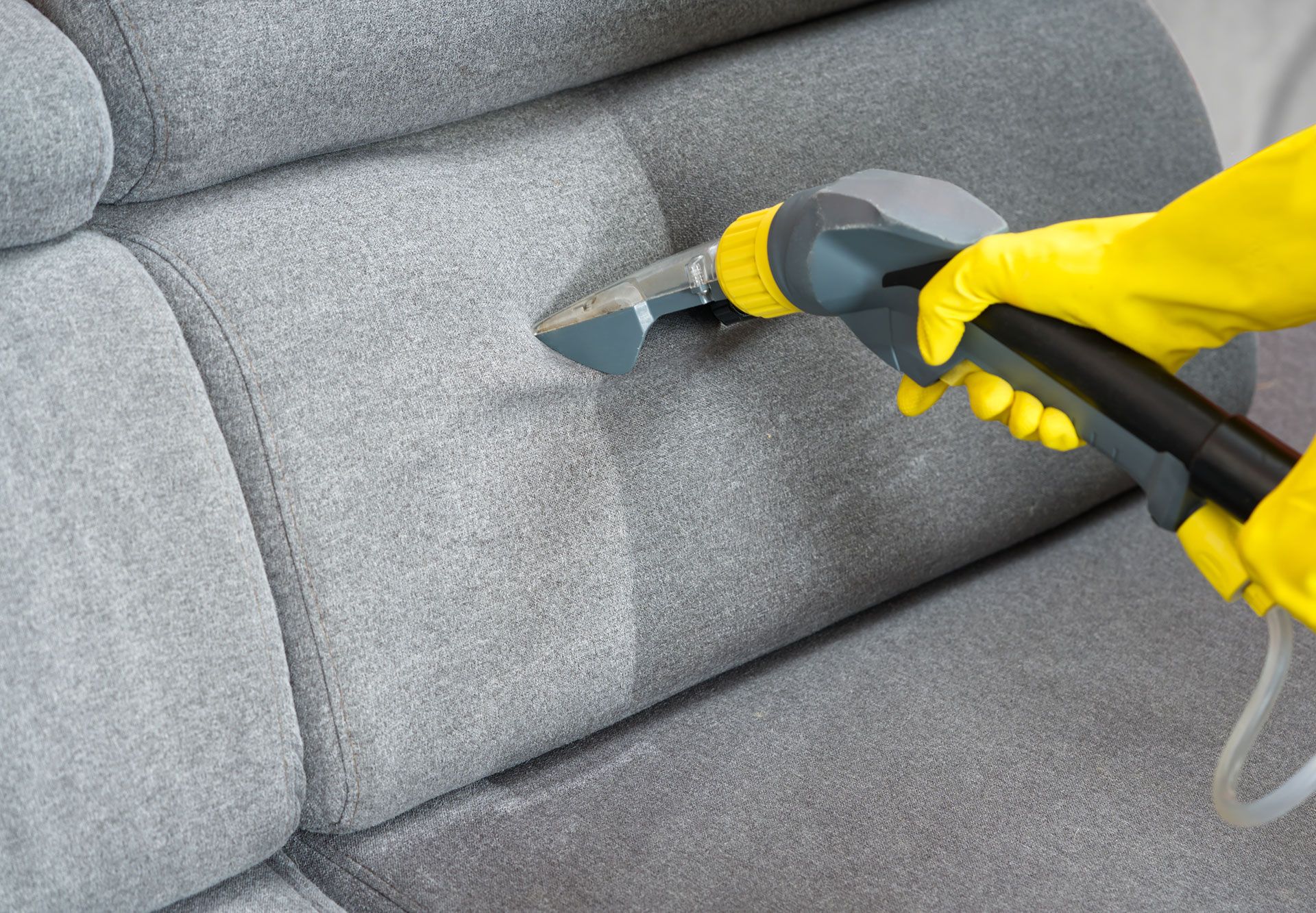Yellow-gloved hand cleaning a gray upholstered sofa with a steam cleaner