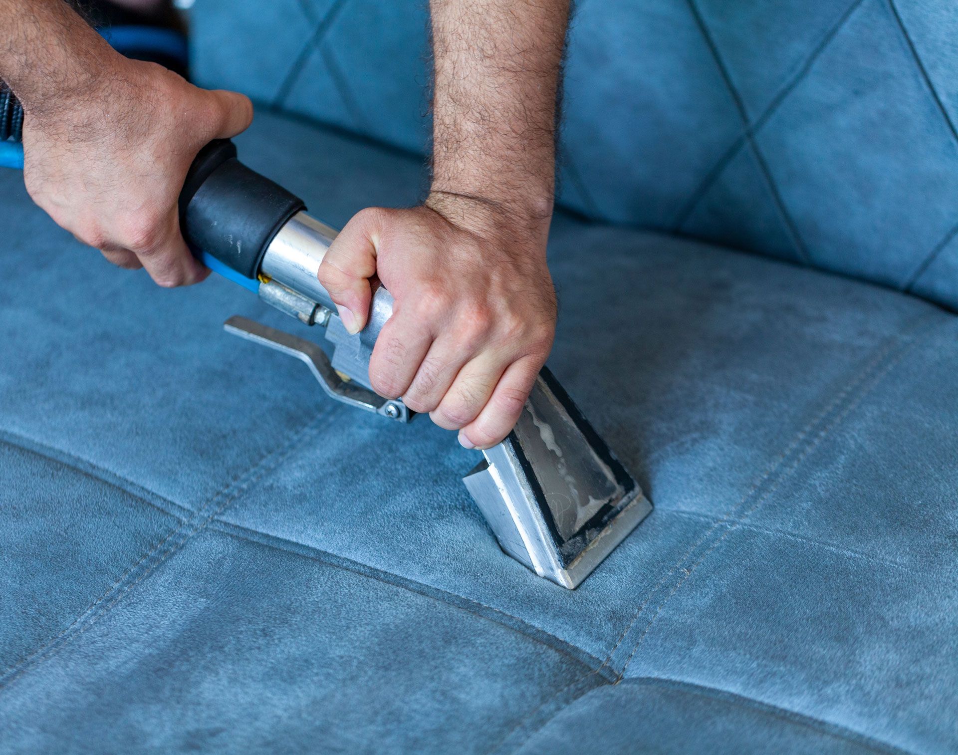 Hands using a handheld upholstery cleaner on a blue fabric sofa.