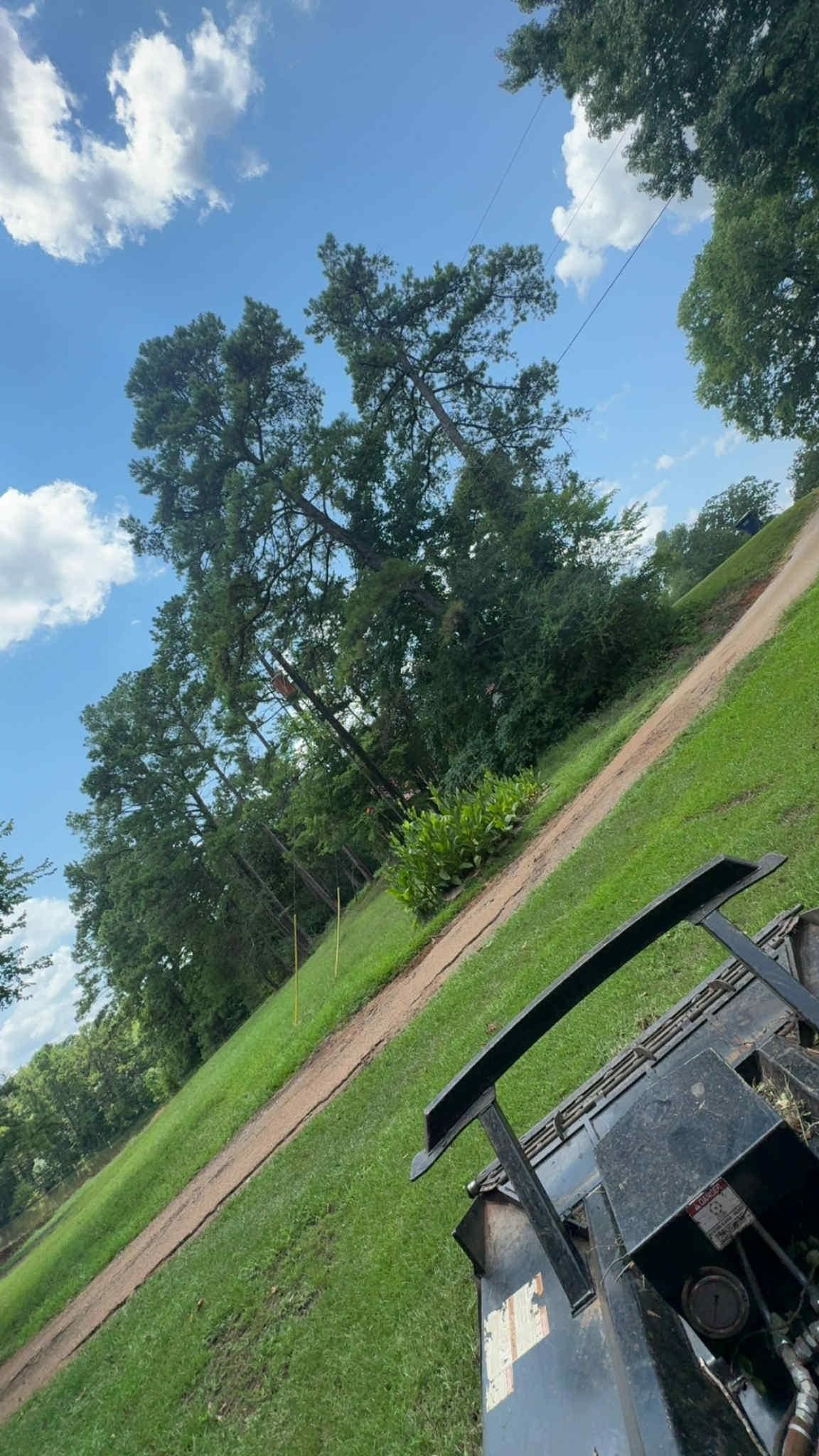 Green grassy hill with dirt path leading toward trees under a blue sky.  Part of a black machine is in the foreground.