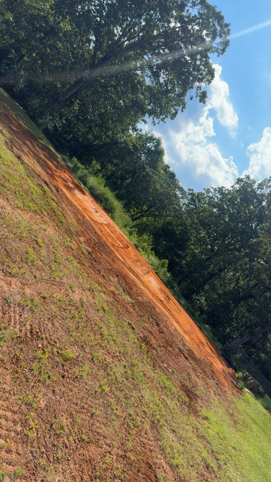 Sloping red dirt path through green grass, bordered by dense trees under a blue sky with clouds.