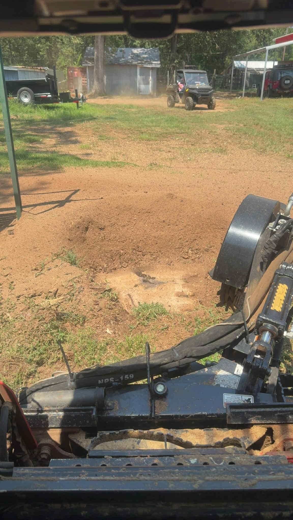 View from inside a machine with a blade. A vehicle moves across a dirt field with other structures in the background.