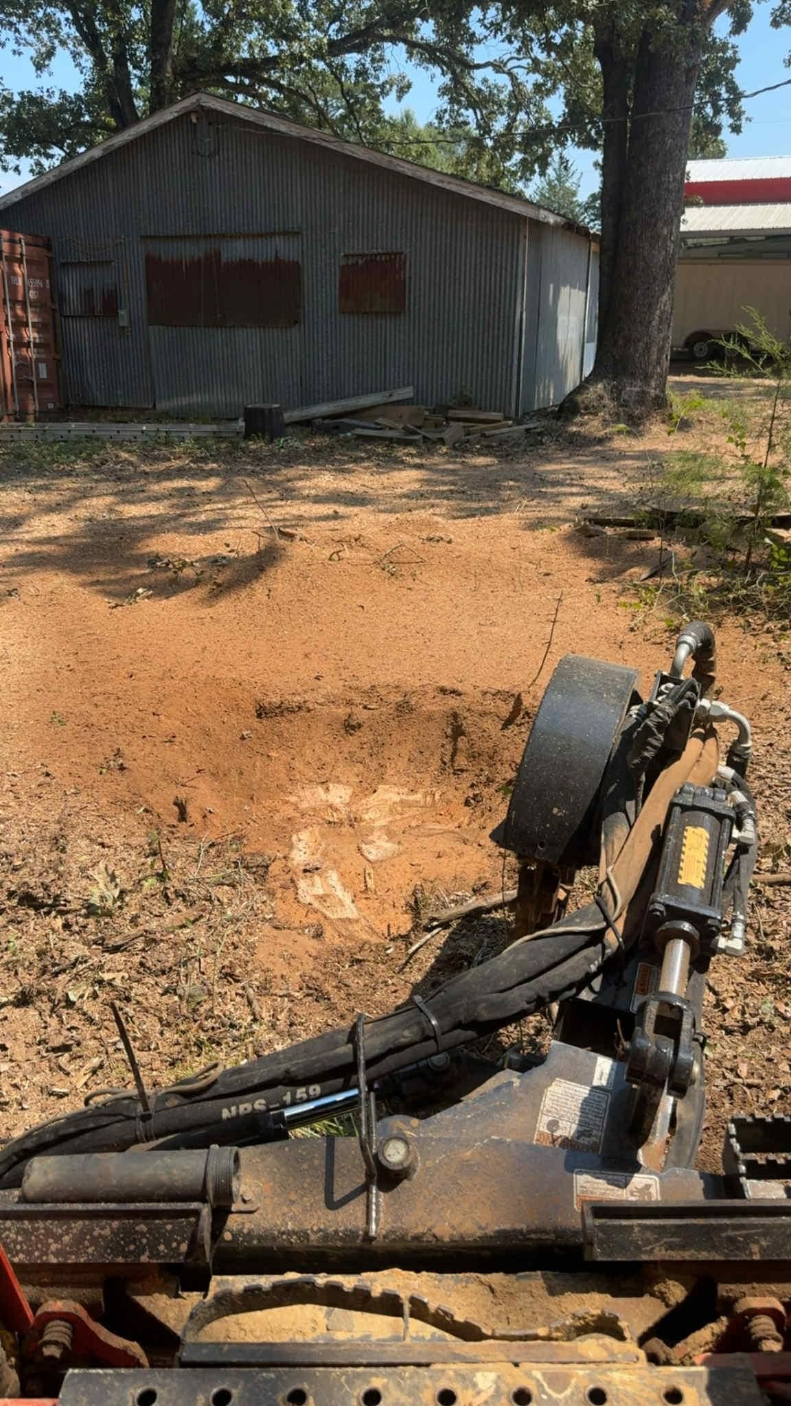 A piece of machinery leveling dirt in front of a weathered metal shed on a sunny day.