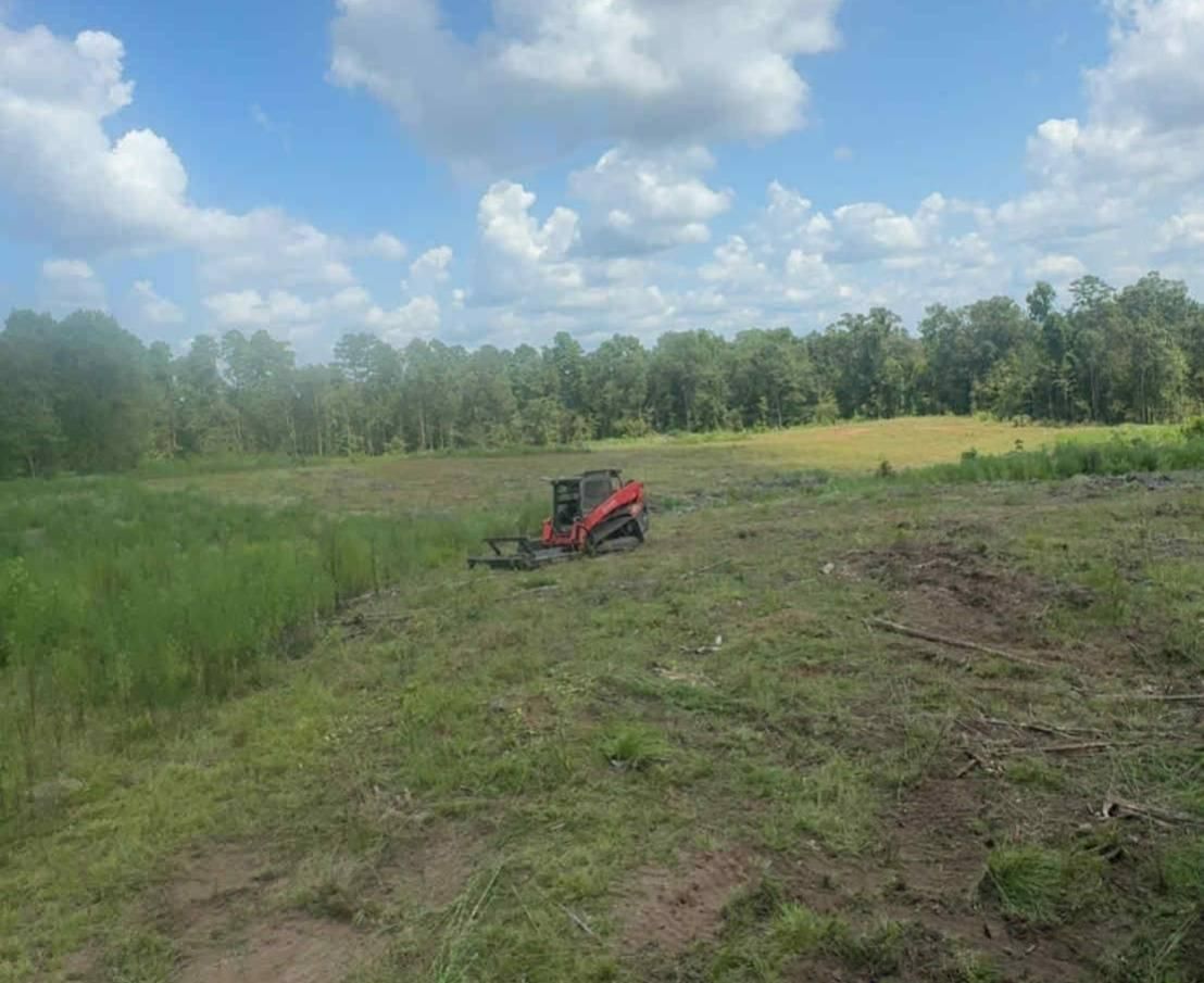 A red mini-skid steer with a mower attachment on a grassy field, trees in the background, blue sky with clouds.