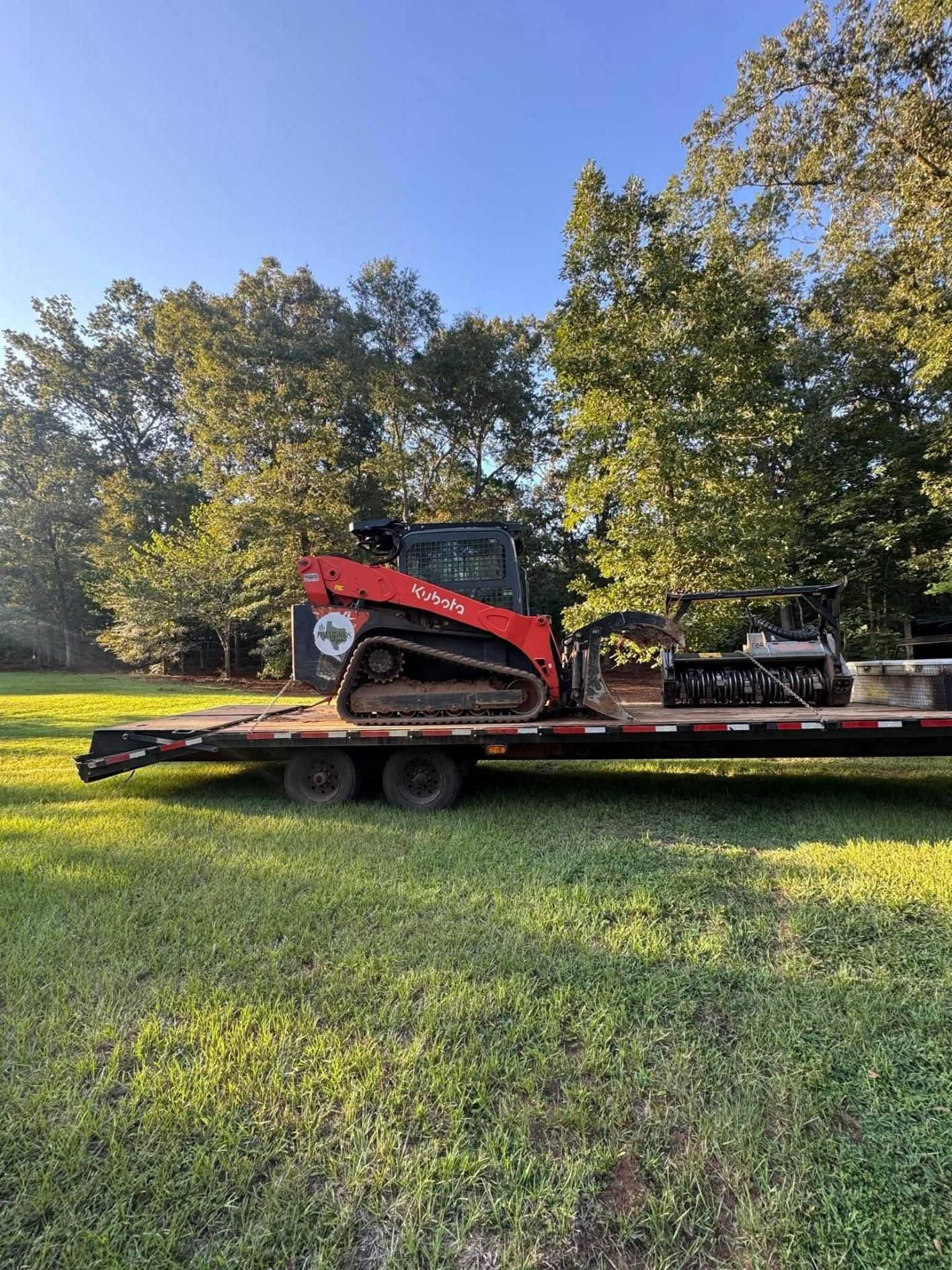 Red skid steer on a trailer in a grassy field, trees in the background under a blue sky.