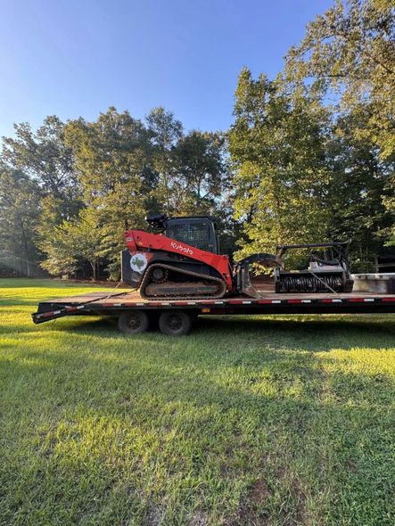 Red track skid steer on a trailer, parked on grass in front of trees under a blue sky.