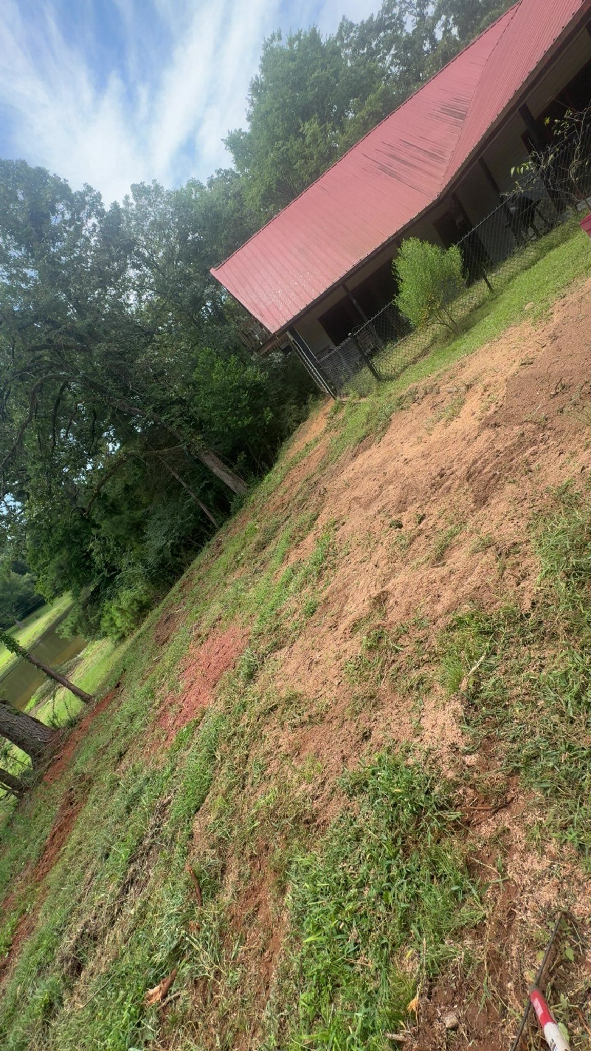 Sloping yard with sparse vegetation, brown soil, and a red-roofed building against a blue sky.