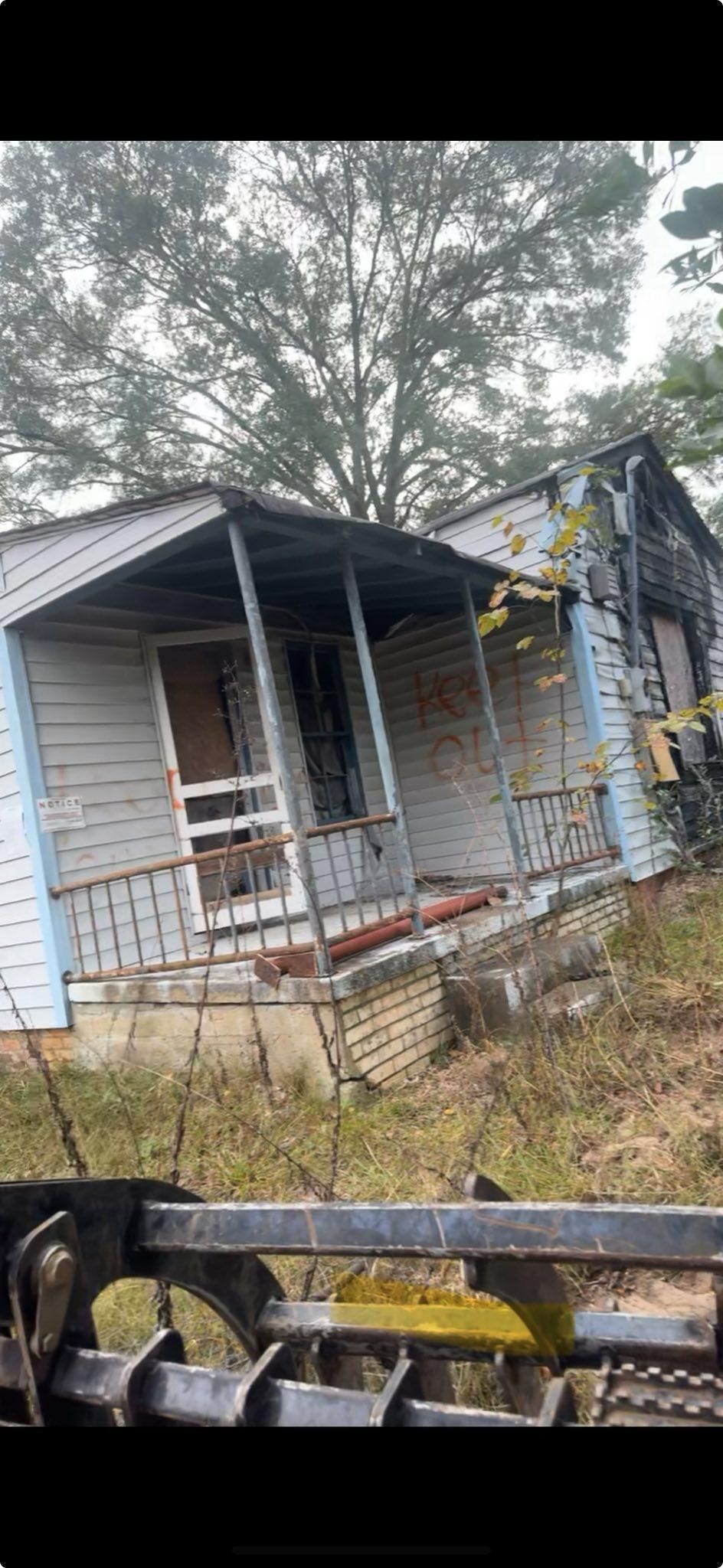Dilapidated, blue-trimmed house with a porch and overgrown weeds, viewed from an earthmoving machine.