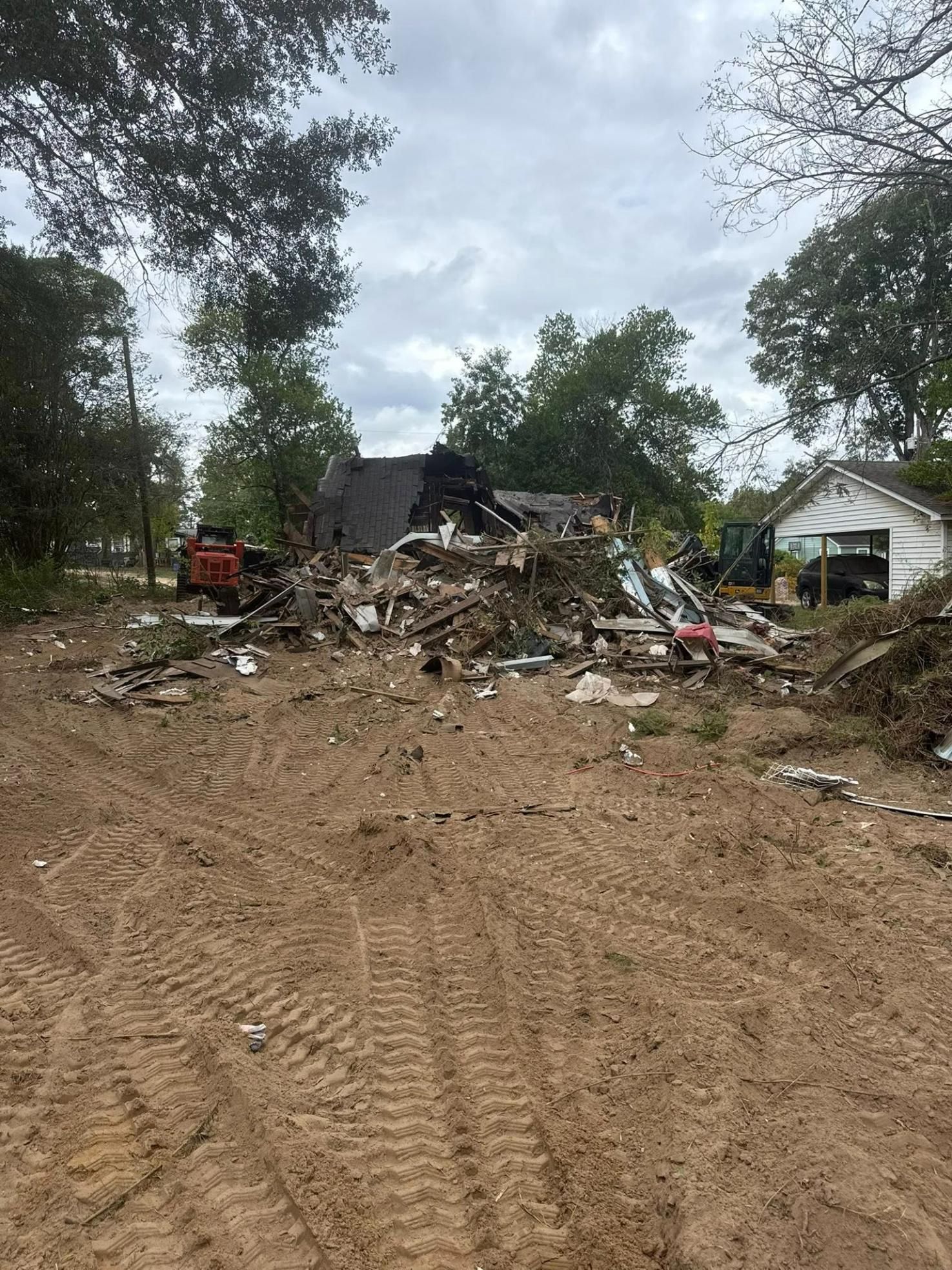 Debris pile from a demolished building in a cleared dirt lot, with trees and a small white building in the background.