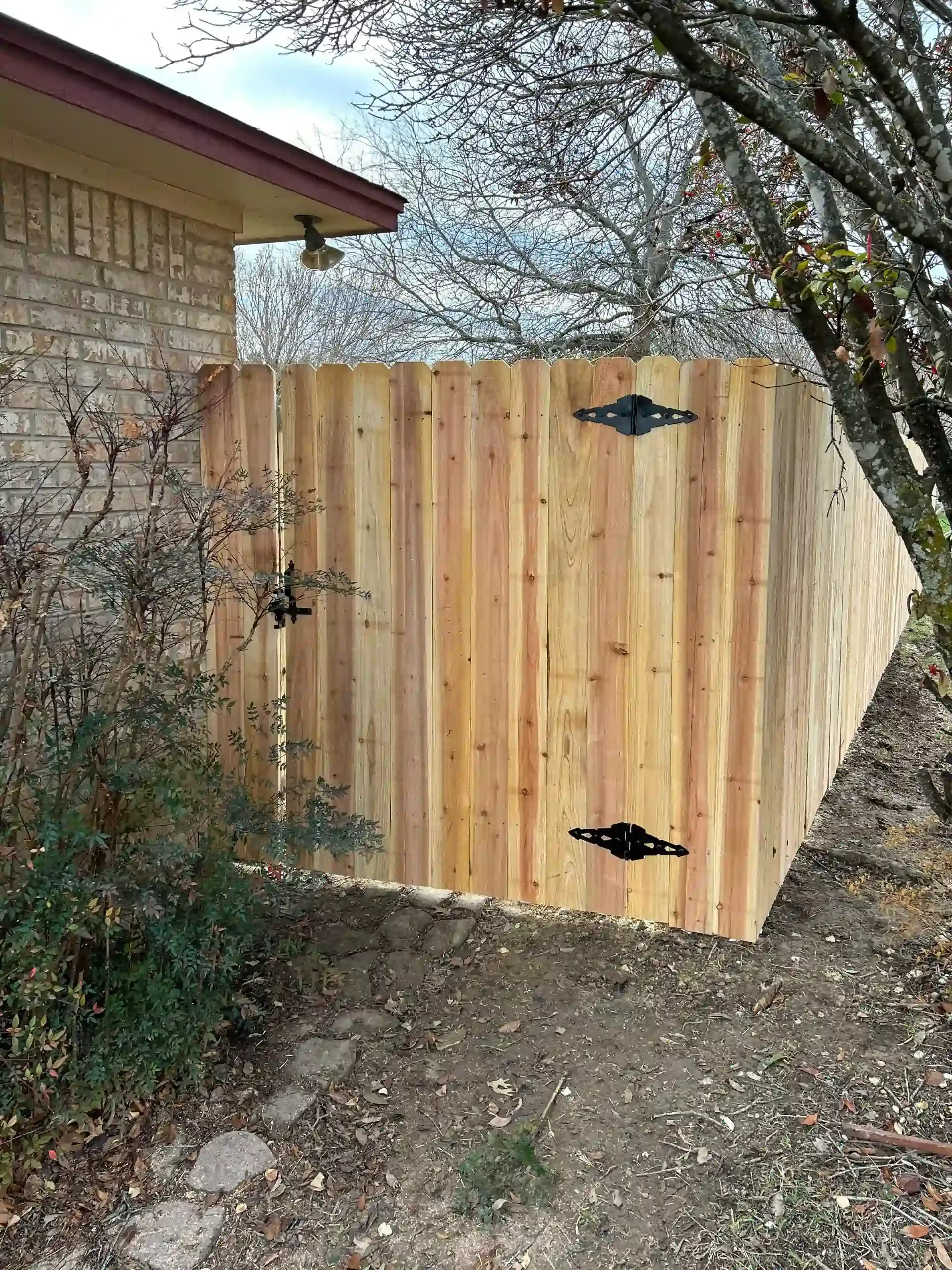 Wooden fence with black hinges against a brick wall and natural surroundings.