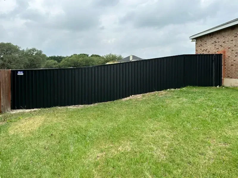 Black corrugated metal fence in a yard with green grass and a brick house.