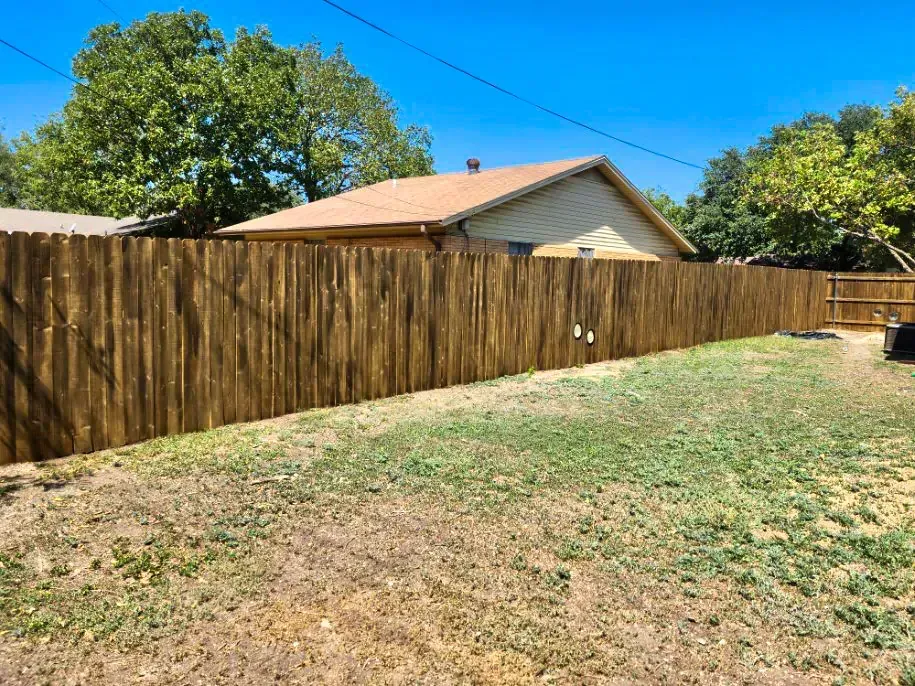 Brown wooden fence surrounds a grassy yard, with a house and trees in the background under a blue sky.