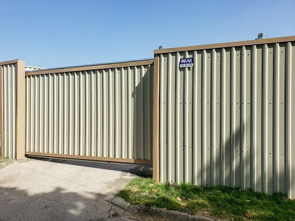 Tan corrugated metal gate and fence on concrete and grass, under a blue sky.