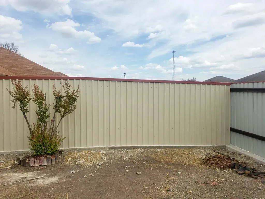 Beige corrugated metal fence in a backyard, with a small bush and gravel.